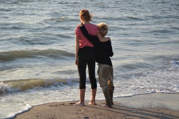 Zwei Kinder von hinten stehen umschlungen vor dem Meer, Junge mit nasser Hose