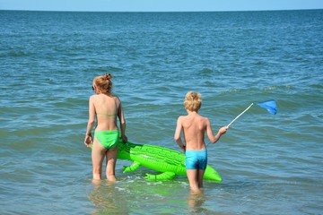 Zwei Kinder  in Badekleidung laufen mit Luftmatratze und Kescher  ins offene Meer