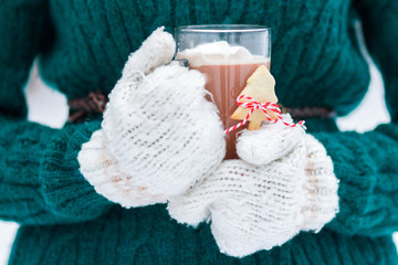 White knitted mittens holding Christmas hot cocoa with melting marshmallows in transparent glass cup