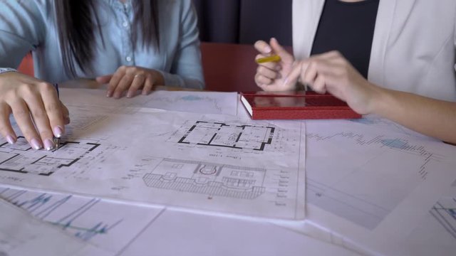 The two realtors works on the report sitting at the table in the co-working room. The papers with house schenes and market situation charts lie on the desk and the professionals outlines the most