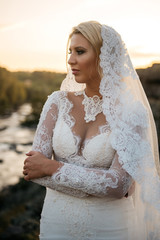 A beautiful bride in a lace chic wedding dress and a long veil, looking thoughtfully into the distance. Sunset, forest and river on the background