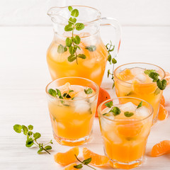 Mandarin cocktail with ice and mint in beautiful glasses and jug, fresh ripe citrus on white wooden background. Sweet orange juice. Close up photography. Selective focus