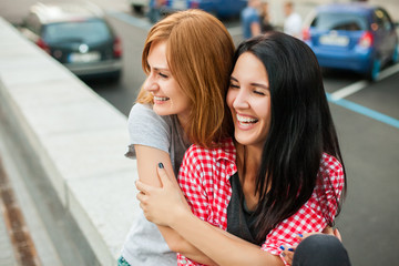 Two atractive young girls hugging, laughting and sitting near road in the street. one brunette girl in red plaid shirt, another redhead girl in gray shirt and blue skirt. concept of sincere friendship