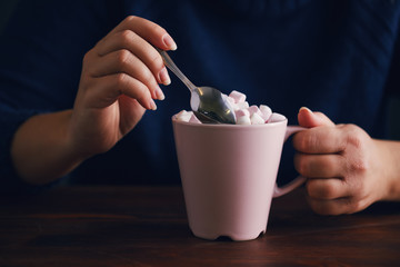 Woman holds pink mug with spoon of hot chocolate and marshmallow