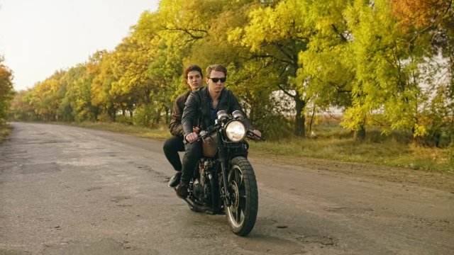 Tracking shot of handsome man in sunglasses riding a motorcycle with his girlfriend behind, traveling together on the asphalt road in forest in autumn