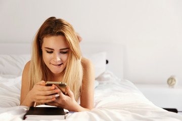 Obraz na płótnie Canvas Beautiful blonde woman looks happy lying on the white bed with a notebook in the morning and checking her phone