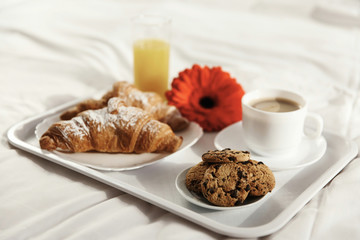 Tray with cup of coffee, glass of juice, red flower, croissants and cookies stands on white bed