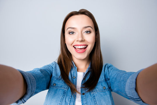  Close up self-portrait of happy  brunette pretty girl with beaming smile in jeans shirt   holding her mobile phone with two hands  standing over grey background