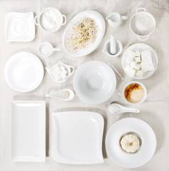 Big set of empty white porcelain plates and other tableware different size and shapes with white breakfast ingredients over white linen tablecloth. Flat lay