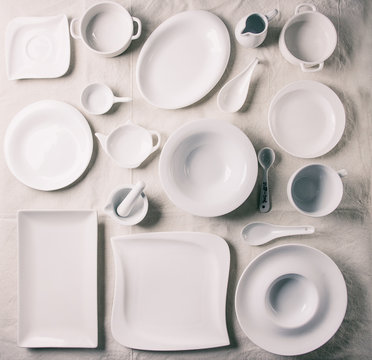 Big set of empty white porcelain plates and other tableware different size and shapes over white linen tablecloth. Flat lay