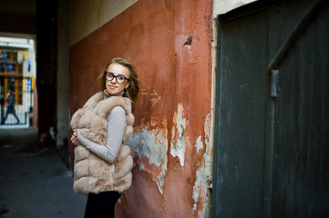 Blonde girl at fur coat and glasses posed against old orange wall.