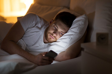 happy young man with smartphone in bed at night - obrazy, fototapety, plakaty