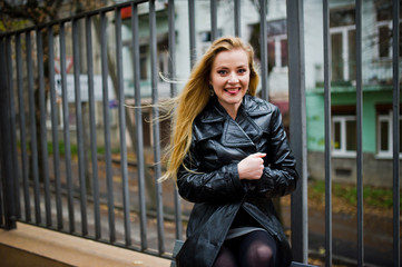 Blonde fashionable girl in long black leather coat posed on bench.
