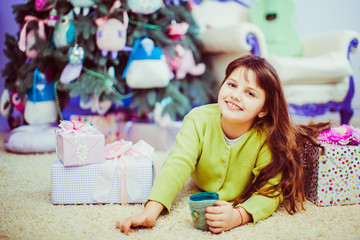 Little girl holds a cup of hot drink lying before a Christmas tree