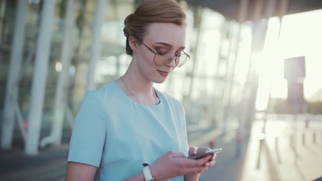 Attractive young woman in stylish glasses smiling charmingly uses her phone and crosses the street. Fashionable look, daily makeup. Modern technologies, active lifestyle, social networks