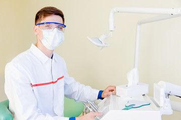 Young dentist student in clinic in workplace with tool and equipment. Man male in white medical gown, mask and glasses.