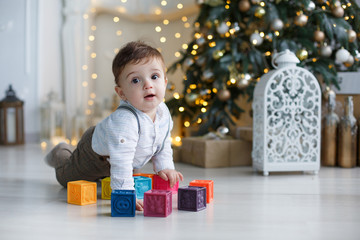 Cute kid with big brown eyes,red hair,dressed in a white striped shirt and brown pants with suspenders,playing at home,on a floor with colored cubes on a green background,beautiful,Christmas tree