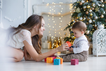 Obraz na płótnie Canvas Happy mother and little son near christmas tree on Christmas. A woman and a little boy are resting in the white bedroom near the Christmas tree. Mother plays with her son waiting for the New Year