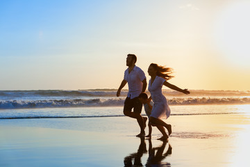 Fototapeta premium Happy full family - father, mother, baby son have fun together, child run with splashes by water pool along sunset sea surf on black sand beach. Travel lifestyle, parents with kids on summer vacation.