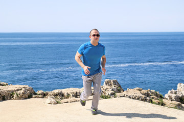 Fit young man jogging on a running trail along seashore. Recreational fitness athlete in sportswear enjoys physical activities on a summer day in a beautiful natural environment. Healthy lifestyle.