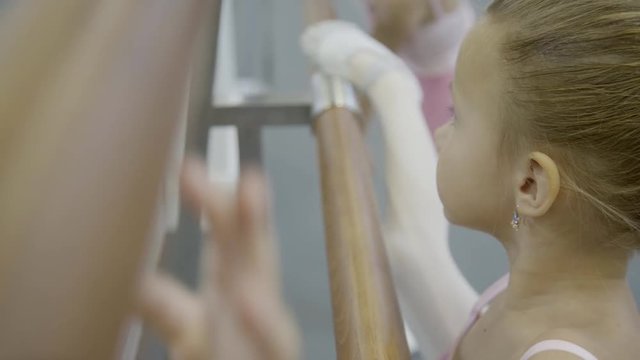 Close-up of little girl is training on stretching legs on wooden crossbar. At ballet school children are engaged in training shoes in dance school.