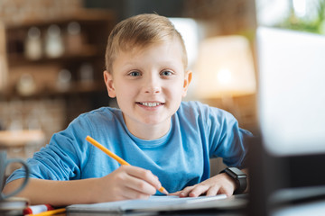 Interesting task. Cheerful delighted intelligent boy sitting at the table and smiling while taking notes