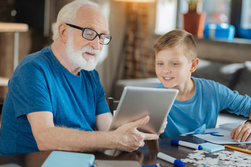 New innovations. Cheerful nice elderly man holding a tablet and looking at its screen while sitting together with his grandson