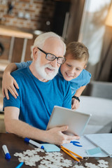 My family. Cheerful positive young boy standing behind his grandfather and hugging him while looking at the tablet screen