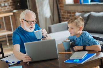 Digital technology. Positive nice aged man holding a laptop and having tea while resting at home with his grandson