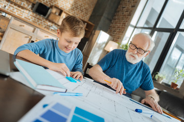 Engineering blueprint. Happy positive delighted boy sitting together with his grandfather and looking at the blueprint while working with him on a project