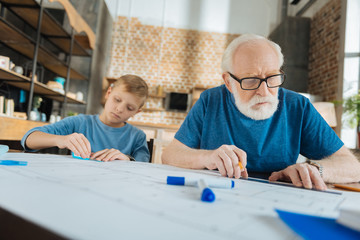 Professional blueprint. Serious smart elderly man sitting with his grandson and doing a drawing while being involved in his job