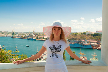 Happy smiling young beautiful blond girl in a white broad-brimmed hat on a background of picturesque seaport