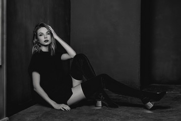 Fototapeta na wymiar Black and white portrait of the young attractive stylish blonde woman in short dress, knee-high boots and black jacket in the minimalistic room with dark walls in photo studio