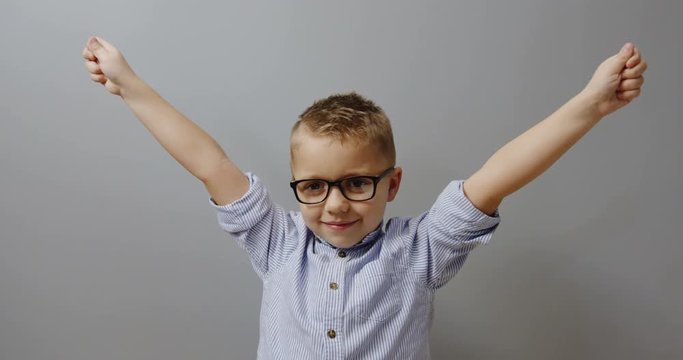 Portrait of the funny cute little boy in the glasses rising hands up like Hurray gesture and looking straight to the camera. on the grey wall background Indoor