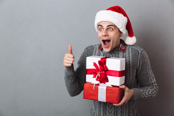 Shocked man in sweater and christmas hat holding gifts