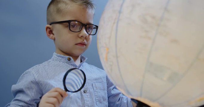 Close up of the nice small boy in glasses standing in front of the globus with a dandruff in his hands and exploring it. Indoor. Portrait shot