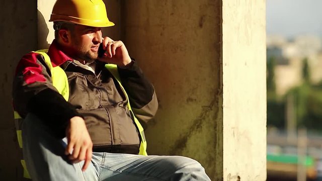 Master builder in yellow hard hat sits on the floor and talks on cell phone. Worker talks on smartphone at project site