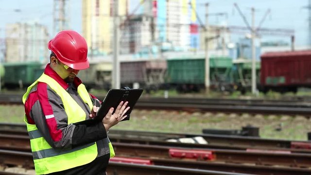 Railway worker with tablet pc at freight terminal. Railway employee makes notes in his tablet pc. Railwayman in uniform and red hard hat works with tablet computer