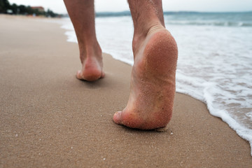 Caucasian man walks on a sand beach. Close up of male feet and golden sand and ocean water. Summer activity and vacation holiday concept