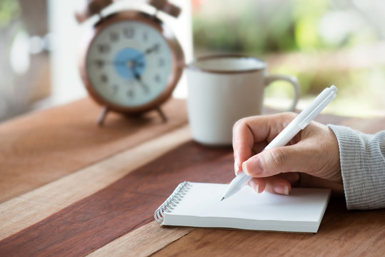 Paper note.Woman hand writing note with white pen on wooden table early in the morning ,blurred alarm clock and  coffee cup in background..