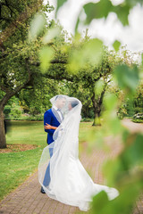 The young happy beautiful newlyweds standing in embrace in the park under bridal veil and looking...