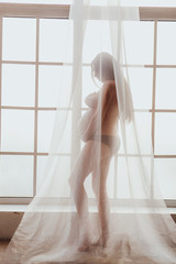Beautiful young mom standing in front of window covered with chiffon curtain.