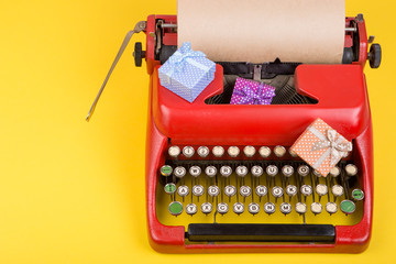 Holidays concept - red typewriter with blank craft paper, gift boxes on yellow background