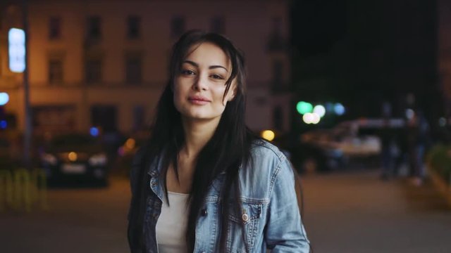 Stylish young brunette in jeans jacket walking down the night city street, looking right towards the camera, smiling seductively. Confident, female beauty, seduction. Having fun