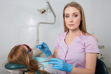 Fototapeta premium Young dental doctor girl holding a medical instrument against the background of a dental office. In the chair sits a patient