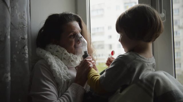 Sick girl with a sore throat makes inhalation with a mask on his face sitting in the window. Son helps his mother to do the inhalation nebulizer