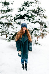 Young beautiful teenager girl in oversized wool coat with long brown beautiful hair standing in winter city park with snowy spruces on background. Fashionable stylish lady with amazaing blue eyes.