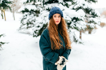 Fototapeta na wymiar Young beautiful teenager girl in oversized wool coat with long brown beautiful hair standing in winter city park with snowy spruces on background. Fashionable stylish lady with amazaing blue eyes.