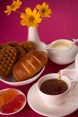 On a dark red background with teapot, Cup with saucer and napkin, sweets, jam, yellow flowers. Tea poured into a Cup. Breakfast. White dishware on Burgundy background. White Cup of tea.