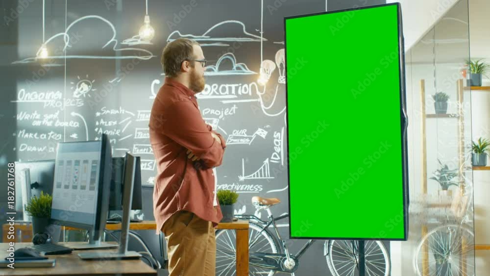 Poster Male App Developer Looks at Interactive Whiteboard with Mock-up Green Screen. He's Thinking on a Problem Solution. In the Background Creative Office with Chalkboard. 